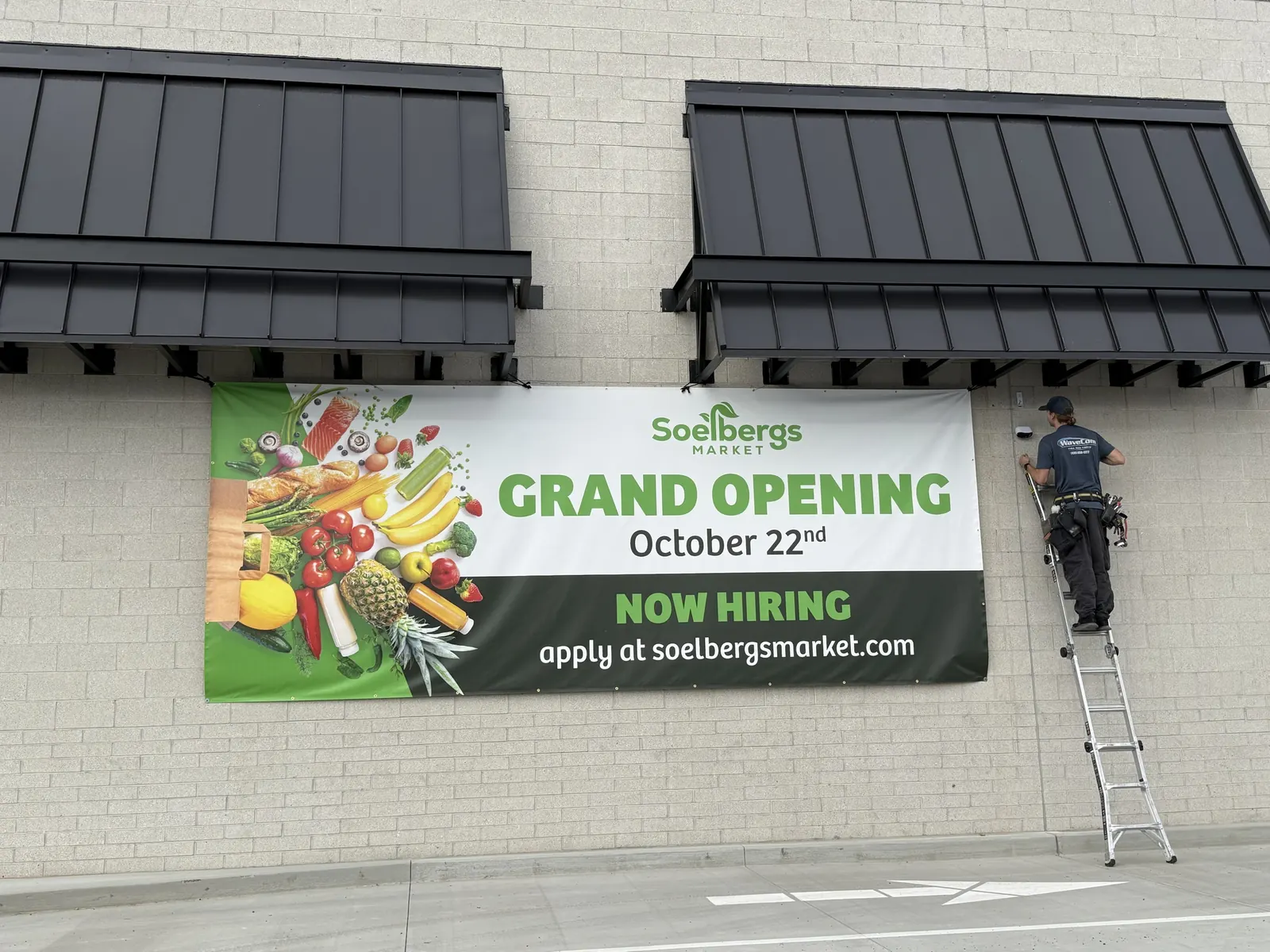 A worker on a ladder next to a Soelbergs banner that reads Grand Opening October 22nd, Now Hiring